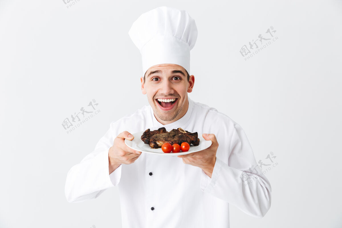 食谱开朗的厨师穿着统一的胡椒粉在一个盘子上煮牛排 隔着白墙热烧烤男性