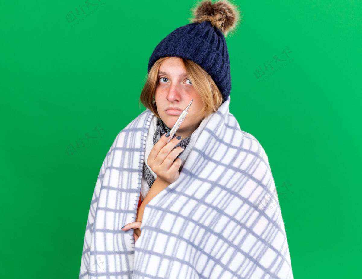 不健康不健康的年轻女子裹着毯子戴着帽子用体温计测量体温患流感发烧站在绿色的墙上女人毯子包裹