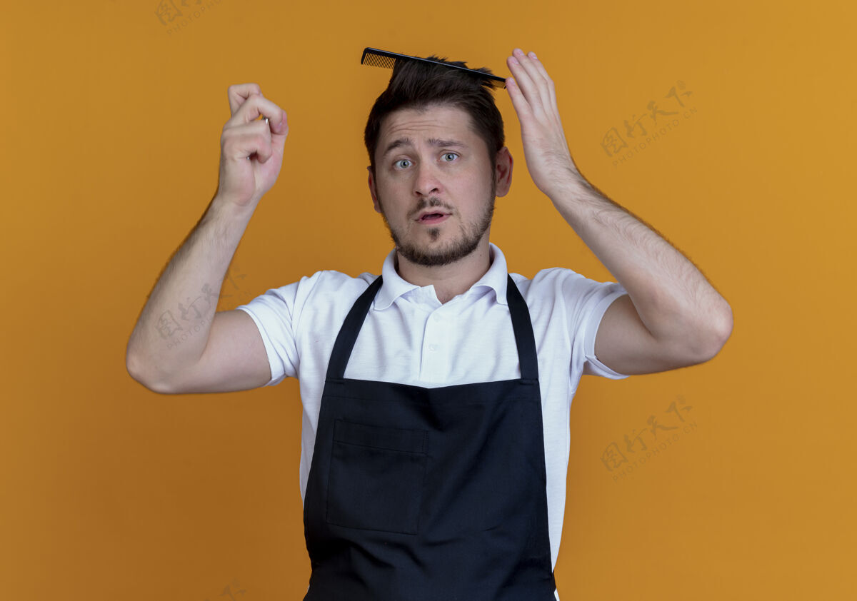 困惑围裙上的理发师拿着毛刷 困惑地站在橙色背景上看着摄像机刷子相机头发
