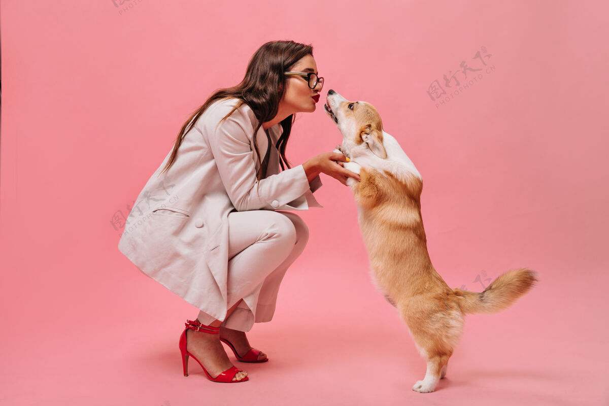 女士穿米色衣服的漂亮女人和粉色背景的狗玩穿着时髦西装和红鞋子的可爱商务女士亲吻柯基犬深色手机狗