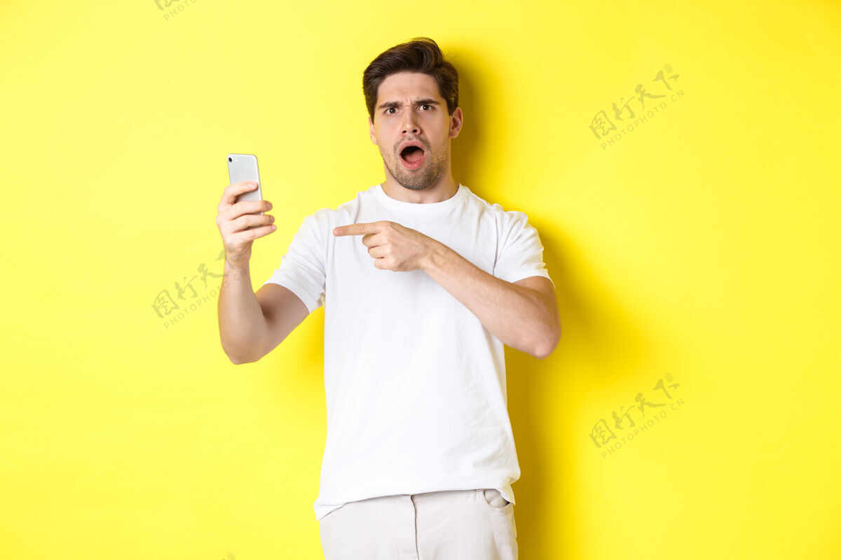 男性困惑的男人用手指着手机屏幕 看不懂什么 站在黄色背景上复制空间成年人移动年轻