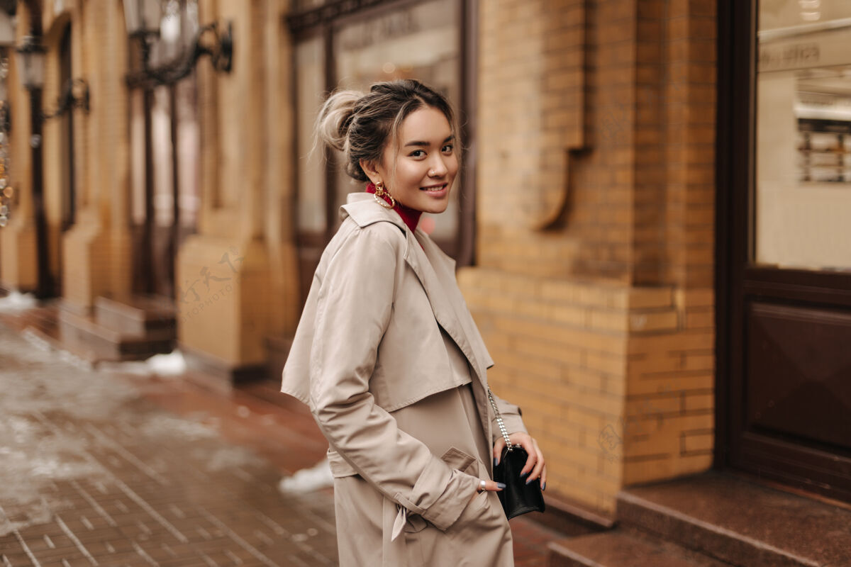 年轻一个微笑的亚洲女人穿着米色的秋衣 拿着一个斜挎包 对着砖砌的老房子摆姿势散步外套阳光