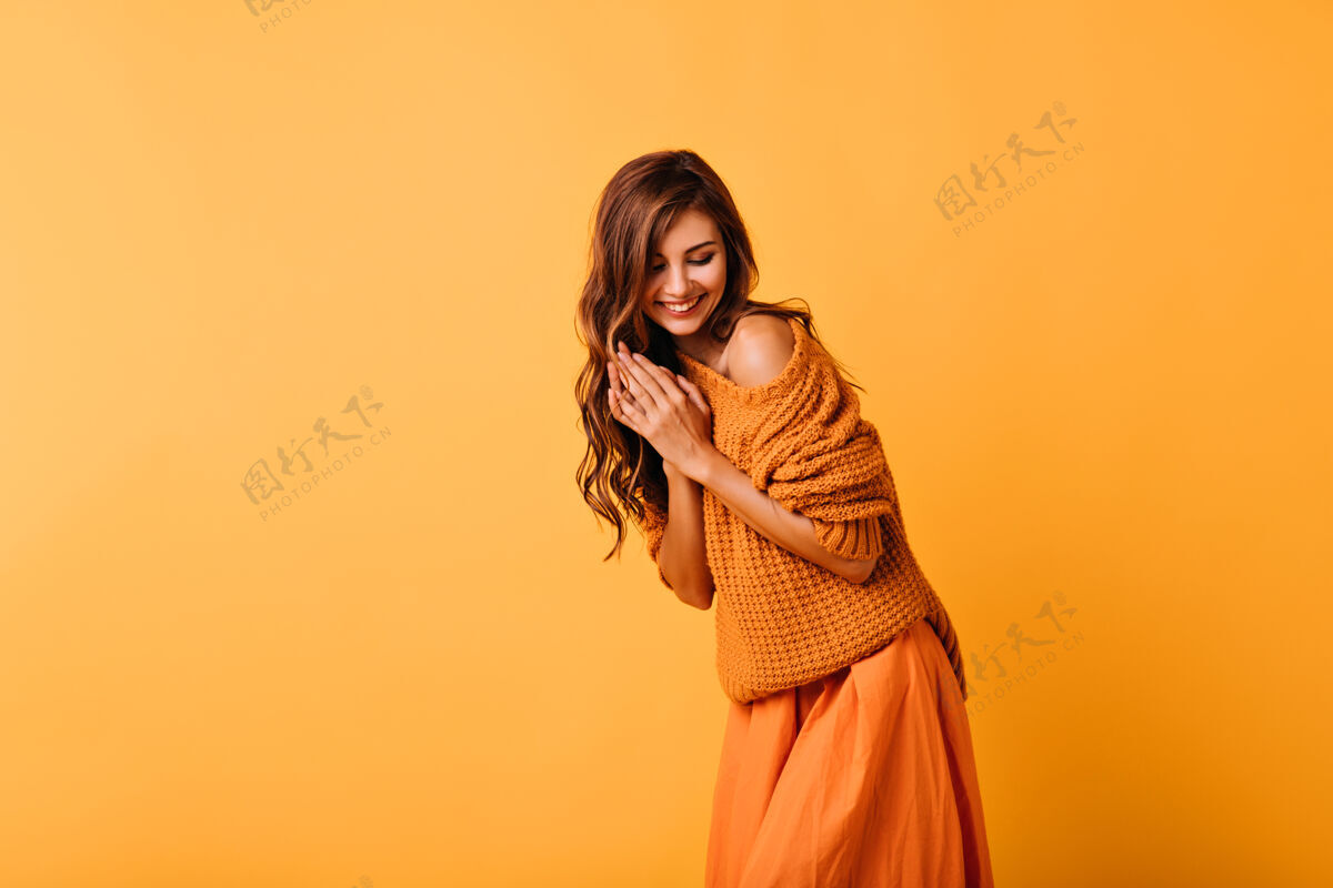 快乐带着波浪发型的浪漫女孩微笑着摆在橙色的脸上穿着时髦毛衣的令人惊叹的白色女士的室内肖像服装有趣放松