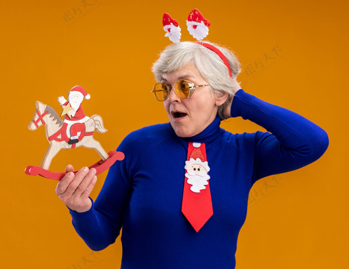 眼镜一位戴着太阳眼镜 戴着圣诞老人头带 打着圣诞老人领带的惊讶的老妇人拿着圣诞老人 望着摇马上的圣诞老人 背景是橙色 有复制空间抱着老人马