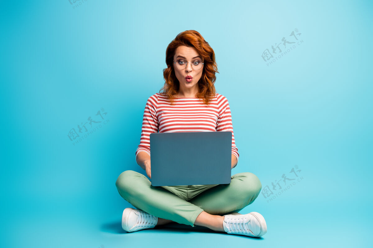 自由职业者惊讶女孩坐着双腿交叉在电脑上工作的全长照片阅读社交媒体工作信息文件印象深刻尖叫哇omg穿时髦的衣服孤立的蓝色阅读休闲地板