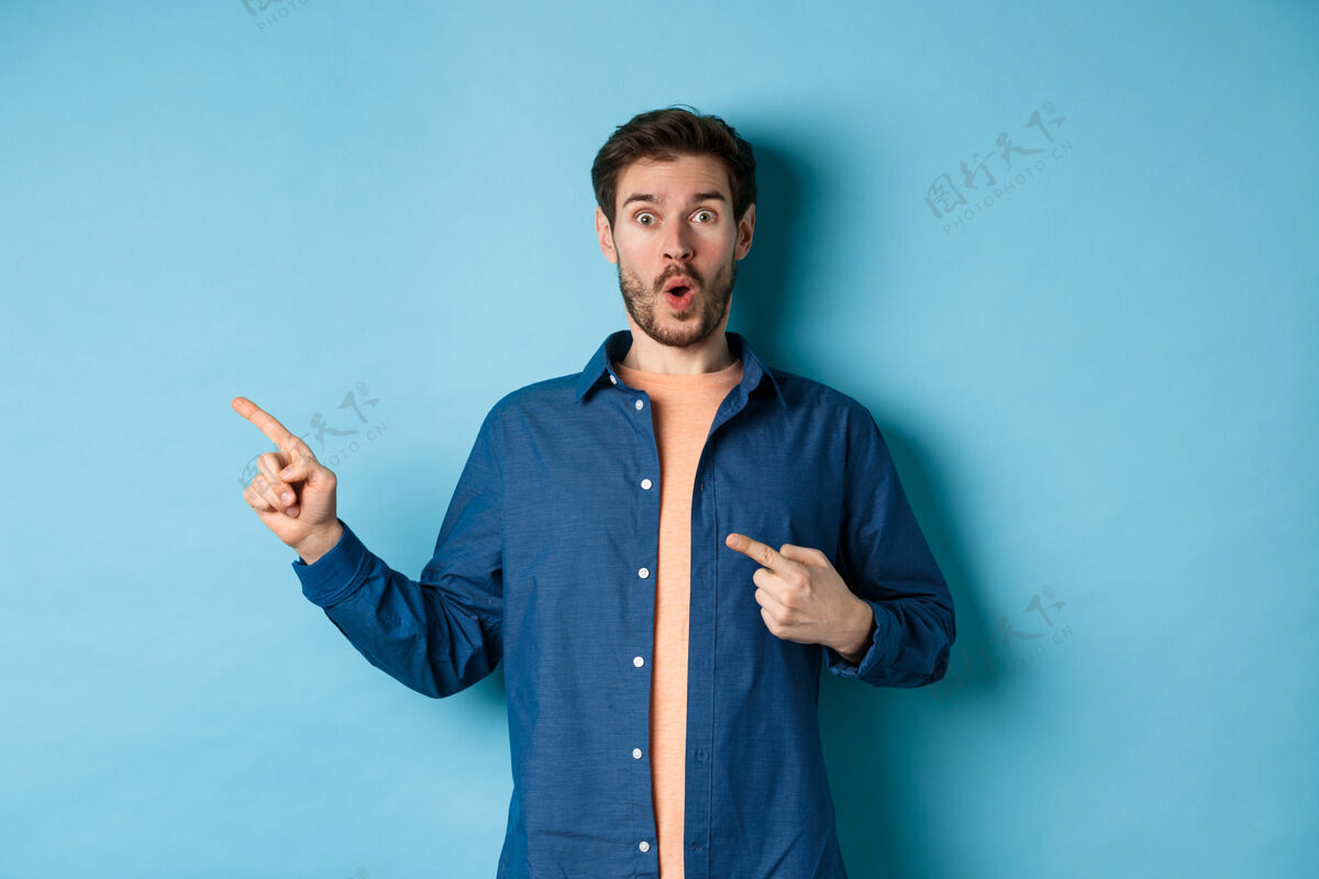 肖像惊讶和惊讶的高加索人在敬畏中喘息 手指指着标志左 显示可怕的宣传片 站在蓝色的背景上指表情站