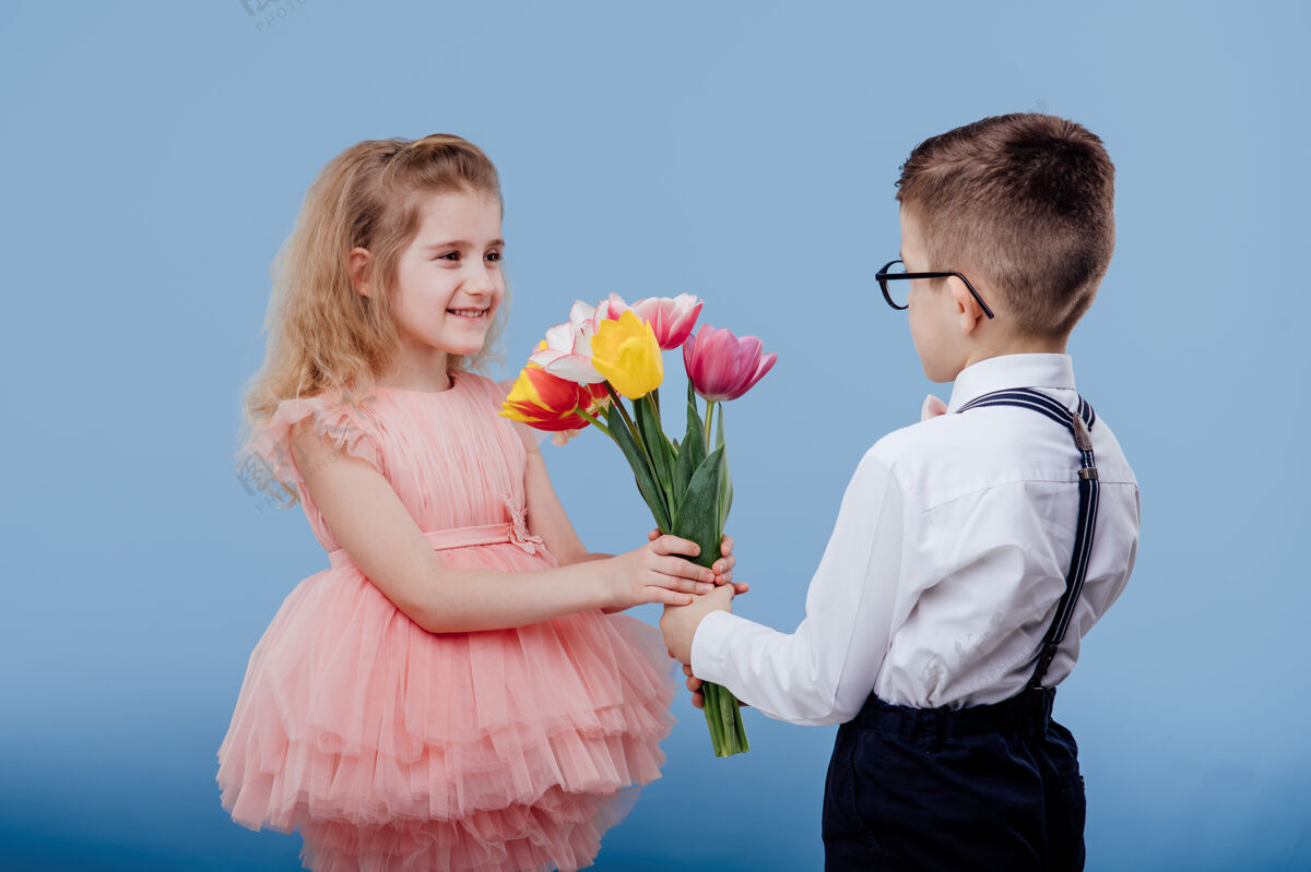 小两个小孩小男孩把花伸向一个穿着粉色裙子的小女孩 隔离在蓝色的墙上情感花微笑
