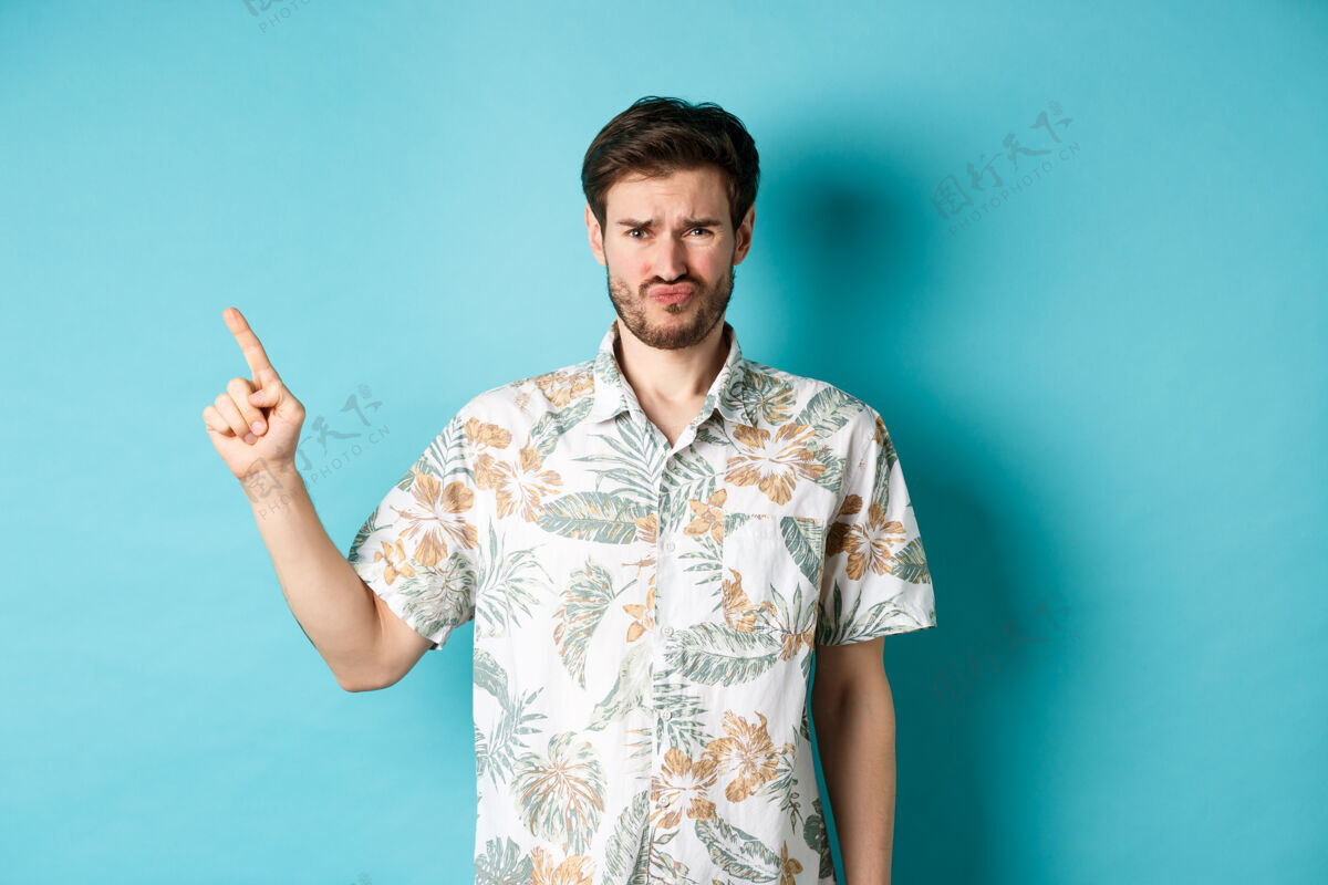 男人失望的游客皱起眉头 用手指着空荡荡的空间 抱怨坏事 穿着蓝色背景的夏威夷衬衫站着胡子指着衬衫
