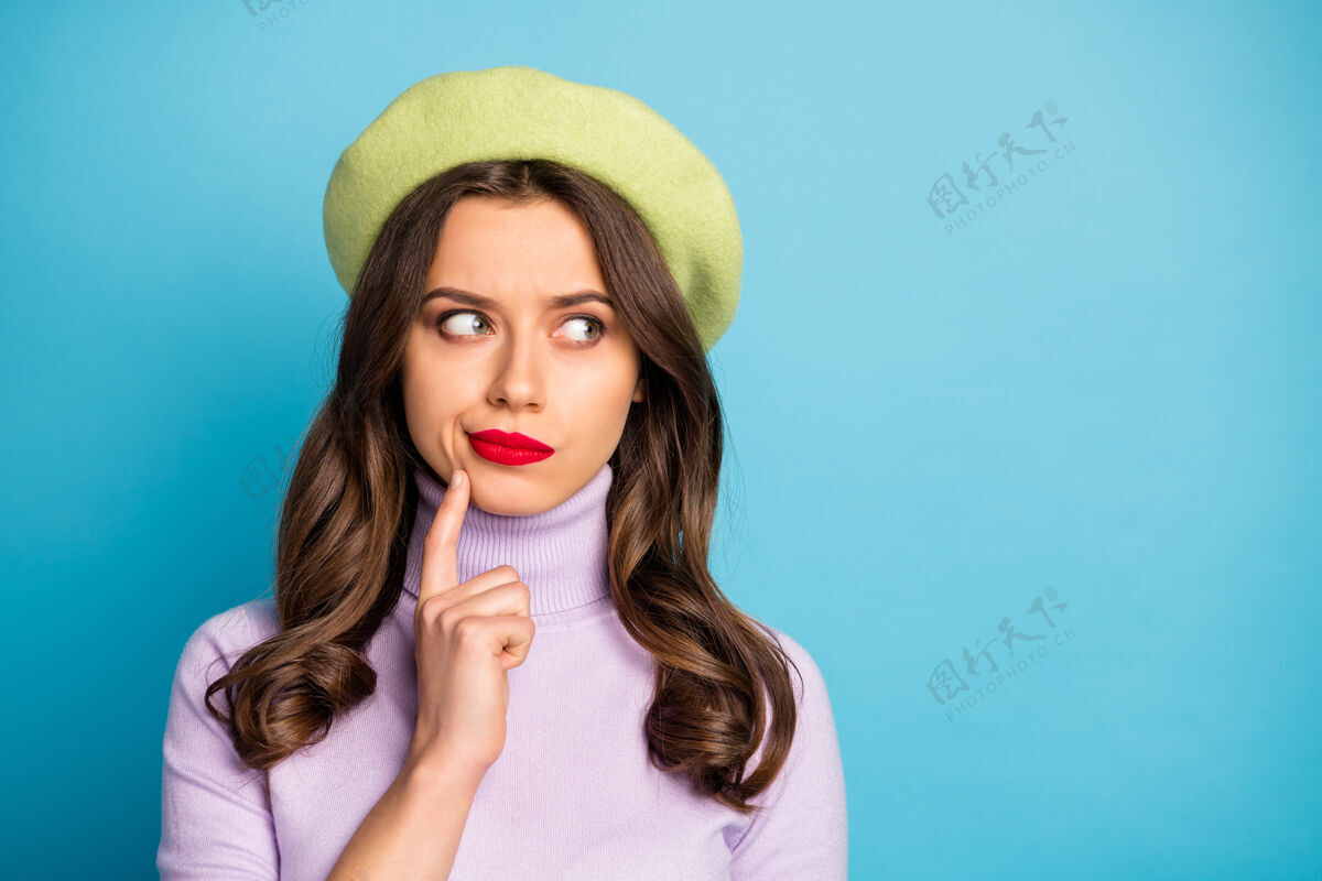 情感美丽旅行者女士的特写照片看边空空手指在下巴上有疑问做决定戴绿色贝雷帽紫色高领毛衣隔离蓝色墙壁穿着法国贝雷帽