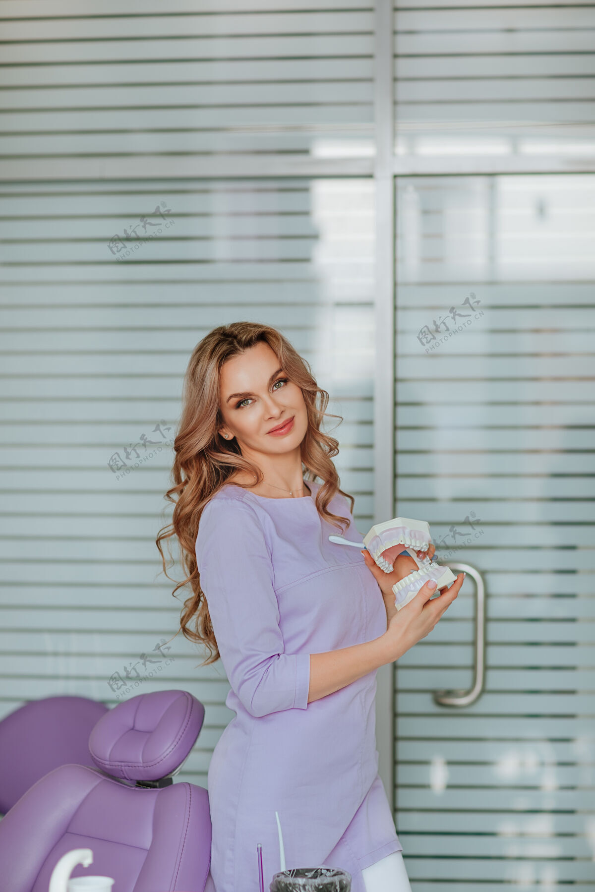 过程年轻迷人的牙科医生的肖像 长卷发 穿着紫色的医疗制服 在橱柜里摆着塑料嘴工具室内医疗