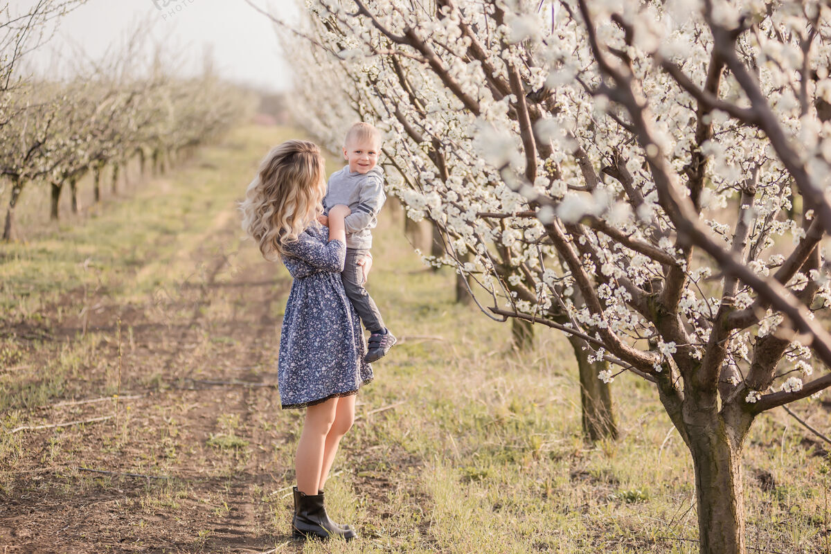 开花年轻的金发母亲带着她的小儿子在盛开的苹果园里手挽着手走着快乐樱桃照顾