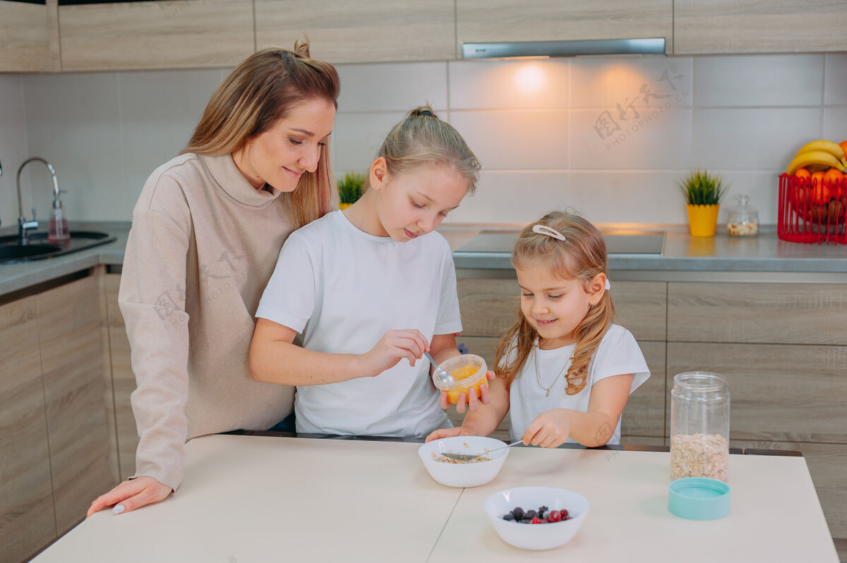 食物一位母亲和她的两个女儿在厨房里给燕麦片加蜂蜜甜室内碗