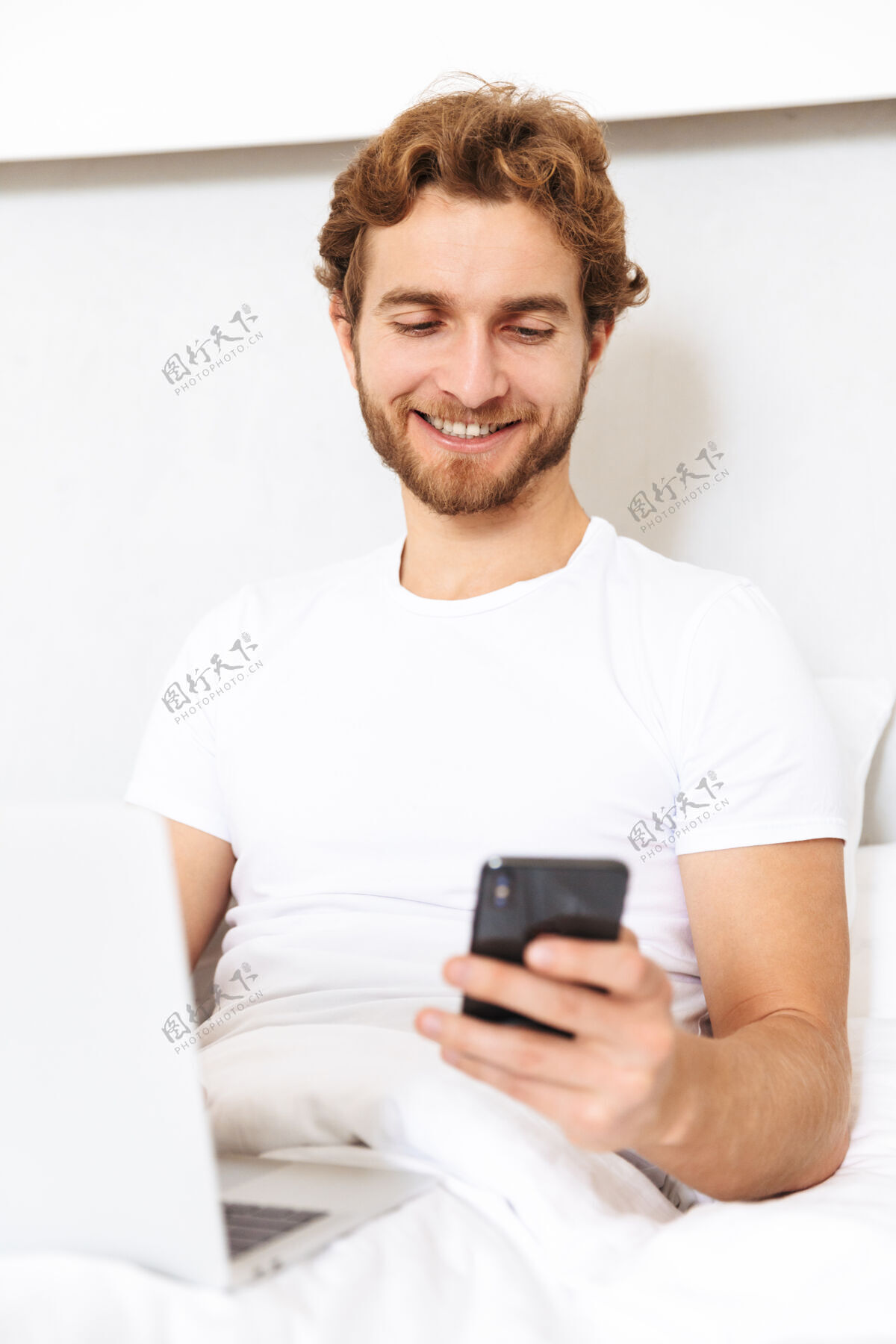 卧室一个英俊的留着胡子的年轻人在家里用笔记本电脑和手机躺在床上室内电话手机
