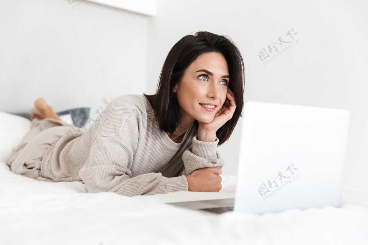 笔记本电脑一张30多岁的轻松女人在明亮的房间里躺在床上用笔记本电脑的照片公寓躺着中年