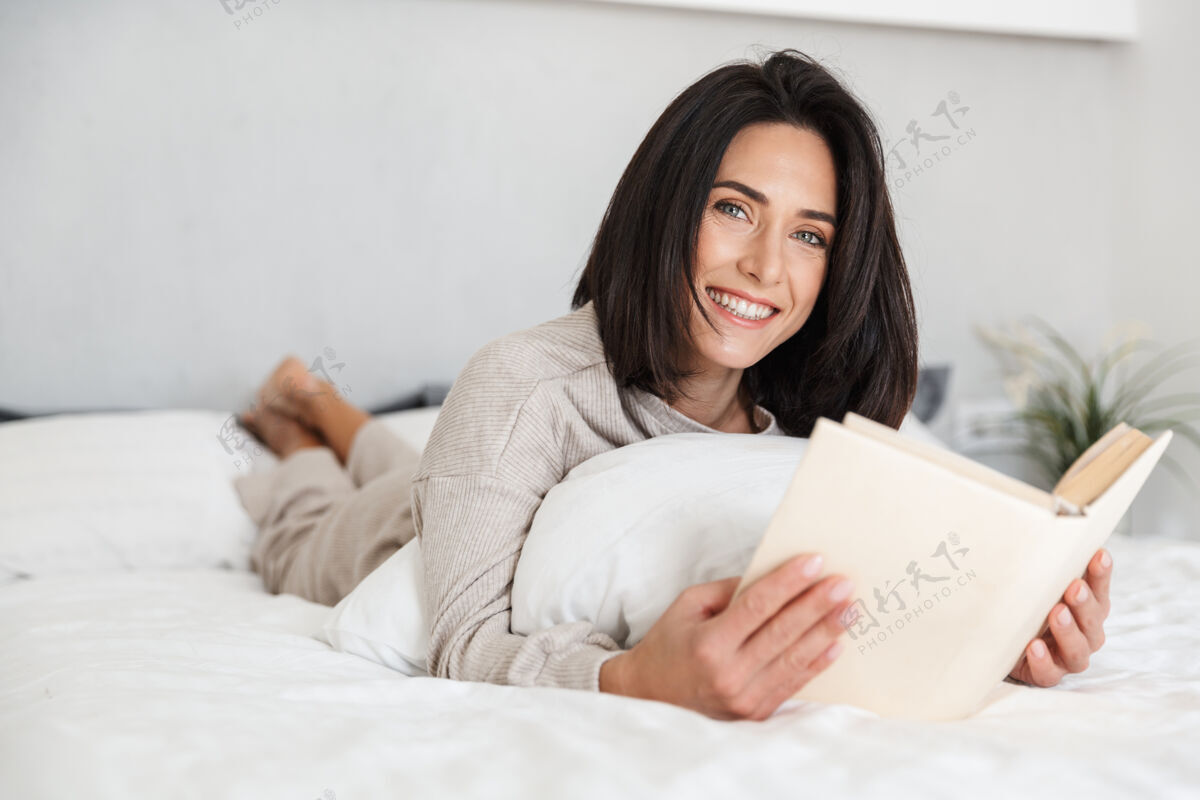 居住图为30多岁的轻松女子一边看书 一边躺在床上 家里铺着白亚麻布中年吸引舒适