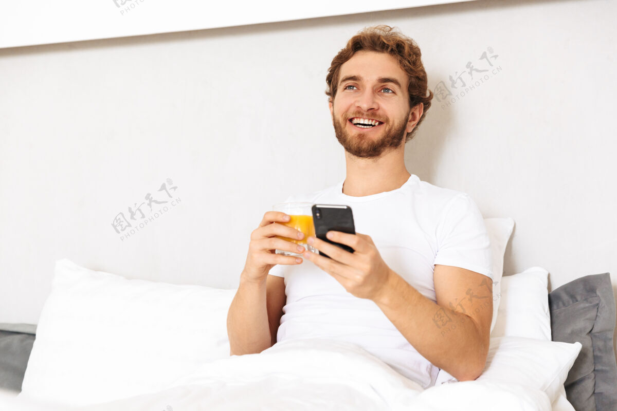 书房一个英俊的留着胡子的年轻人在家里的床上用手机喝果汁短信卧室应用程序