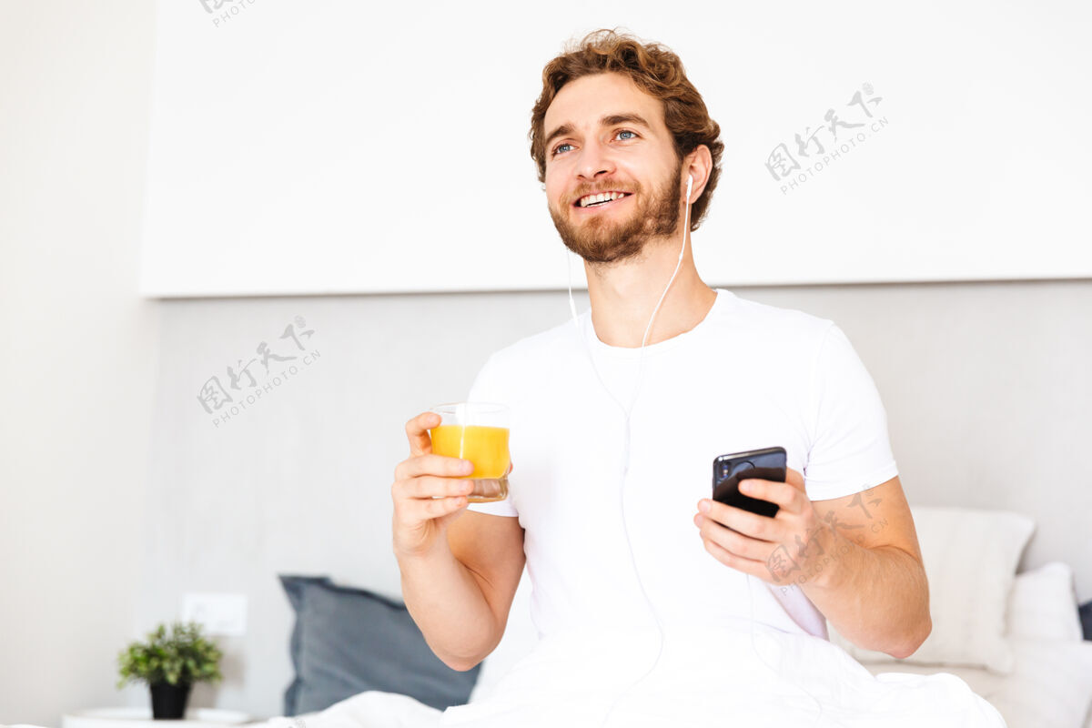 果汁一个留着胡子的帅哥在家用耳机听音乐用手机喝果汁维生素短信听力