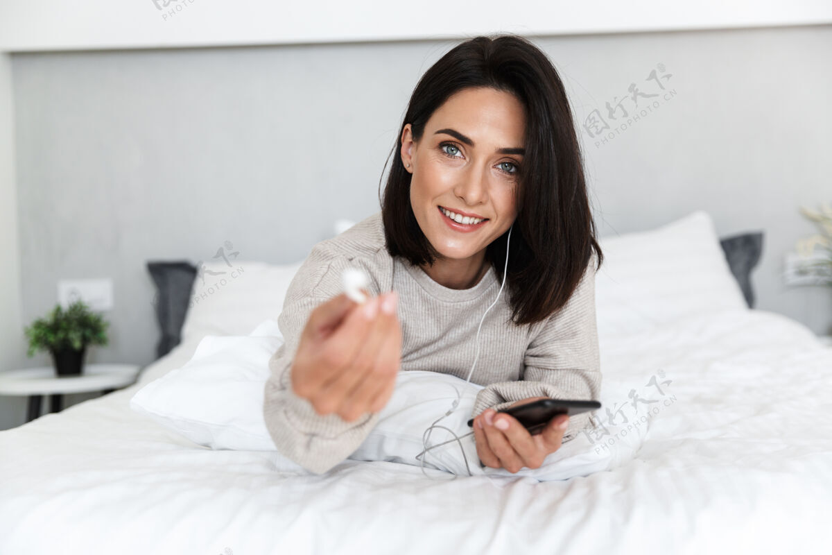 肖像30多岁的欧洲女人戴着耳机拿着手机 躺在明亮的房间里的床上吸引力公寓手机