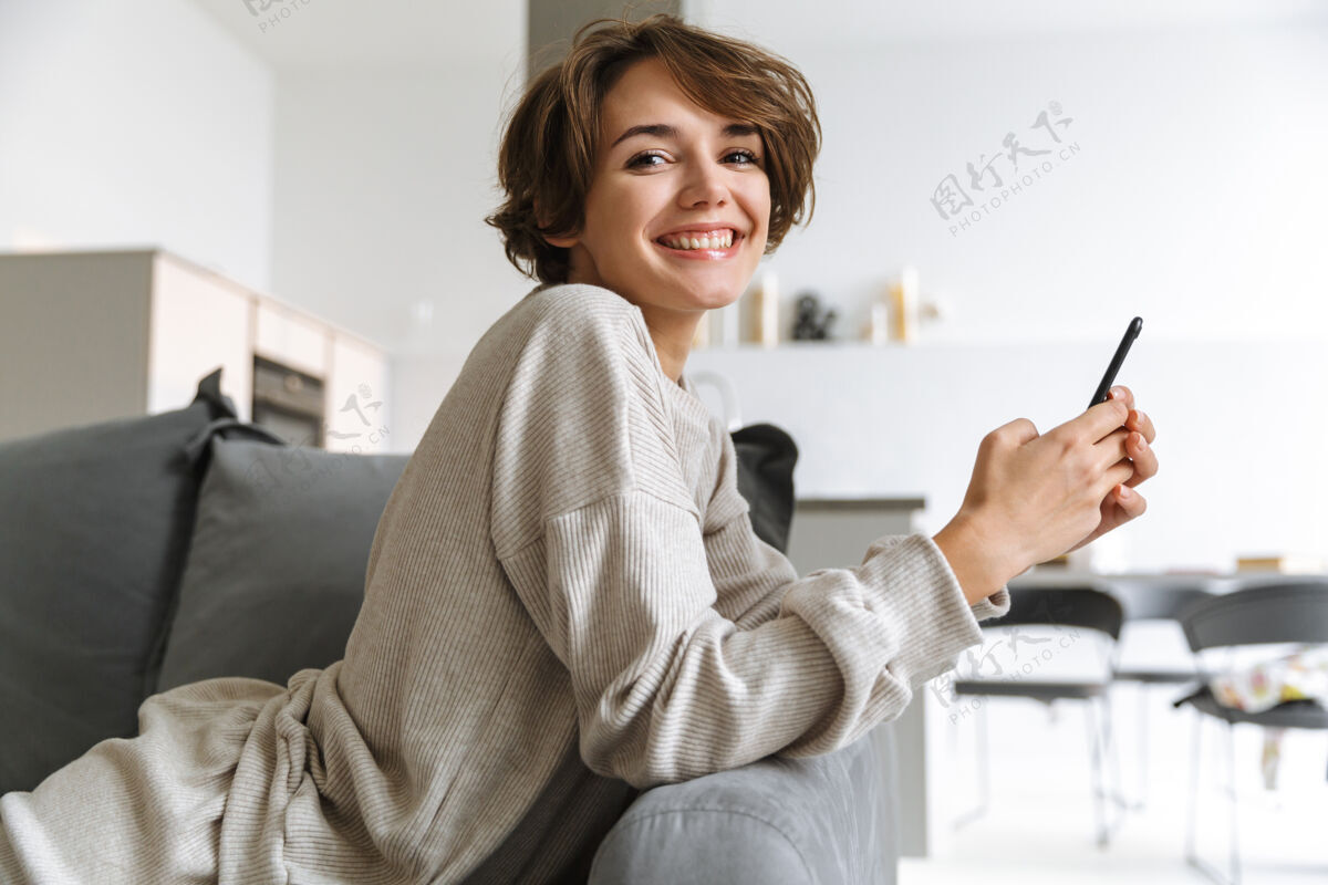 购物快乐的年轻女子坐在家里的沙发上 用手机住所沙发公寓