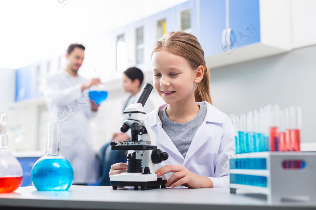 年轻让我看 可爱熟练的小女孩穿着实验服 一边触摸显微镜 一边往下看显微镜科学童年