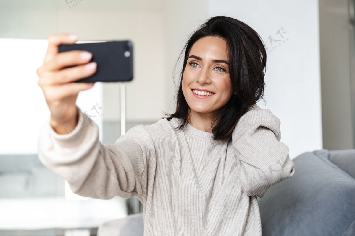 白种人一张30多岁的黑发女人坐在明亮公寓的沙发上用手机自拍的照片小玩意成人设备