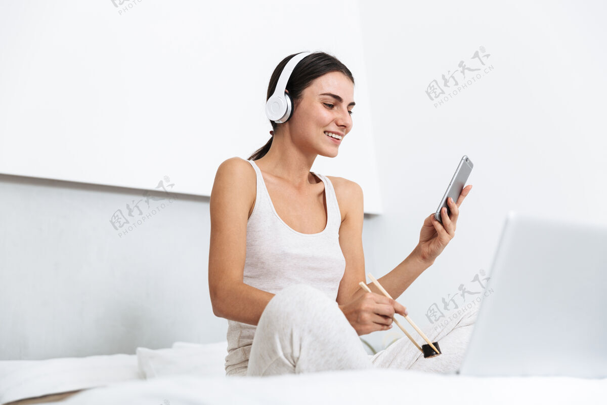年轻美丽的年轻女子在家里的床上放松 戴着耳机听音乐 吃着盘子里的寿司室内耳机新鲜