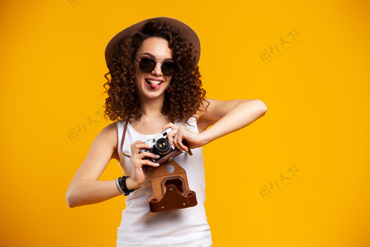 微笑戴着眼镜的笑女青年在复古老式照相机上拍照的肖像 隔离在明亮的黄色上人姿势时尚