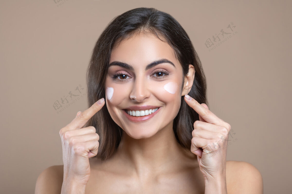 女人面对奶油味的开朗的美丽的年轻女子 带着牙齿般的微笑 手指着她的脸颊使用化妆品健康