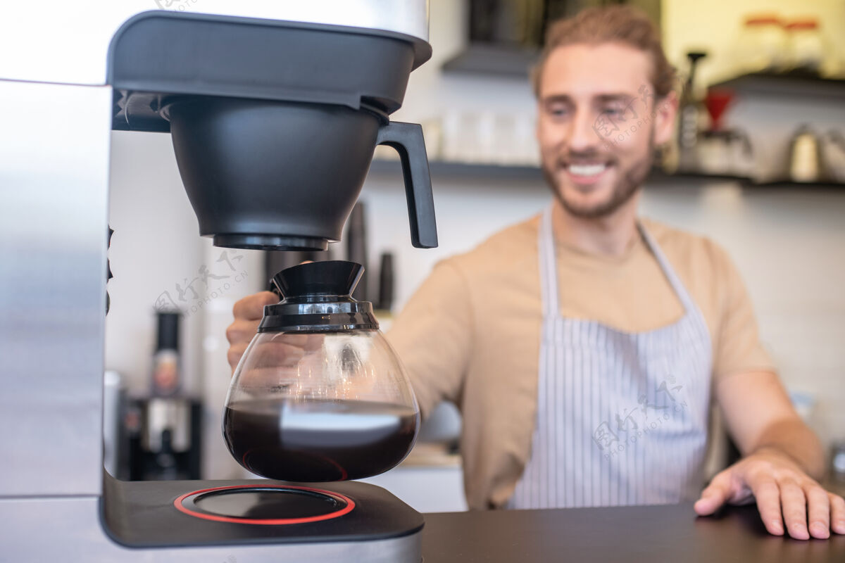 舒适一罐浓缩咖啡快乐的年轻人站在咖啡馆的吧台后面 从咖啡机里拿着一个玻璃壶的咖啡小企业选择性聚焦玻璃壶
