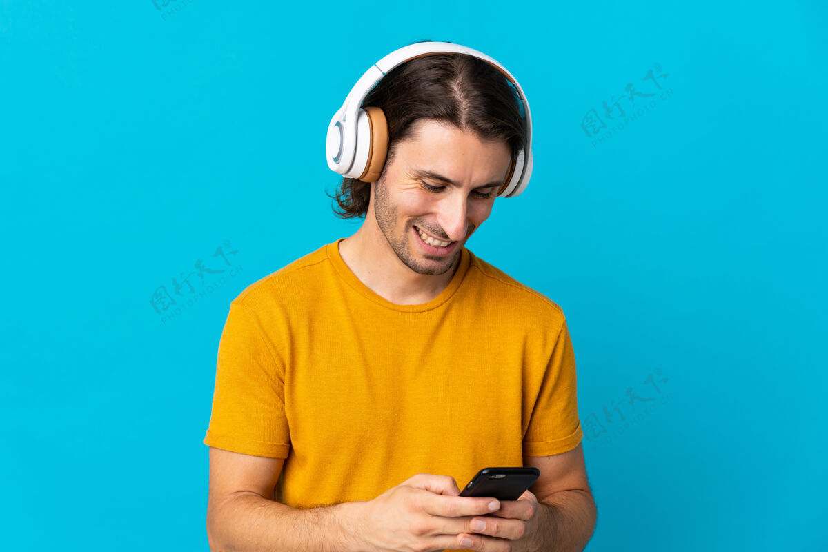 享受年轻帅哥孤零零地站在蓝色的墙上听音乐 望着手机电话休闲瘦身