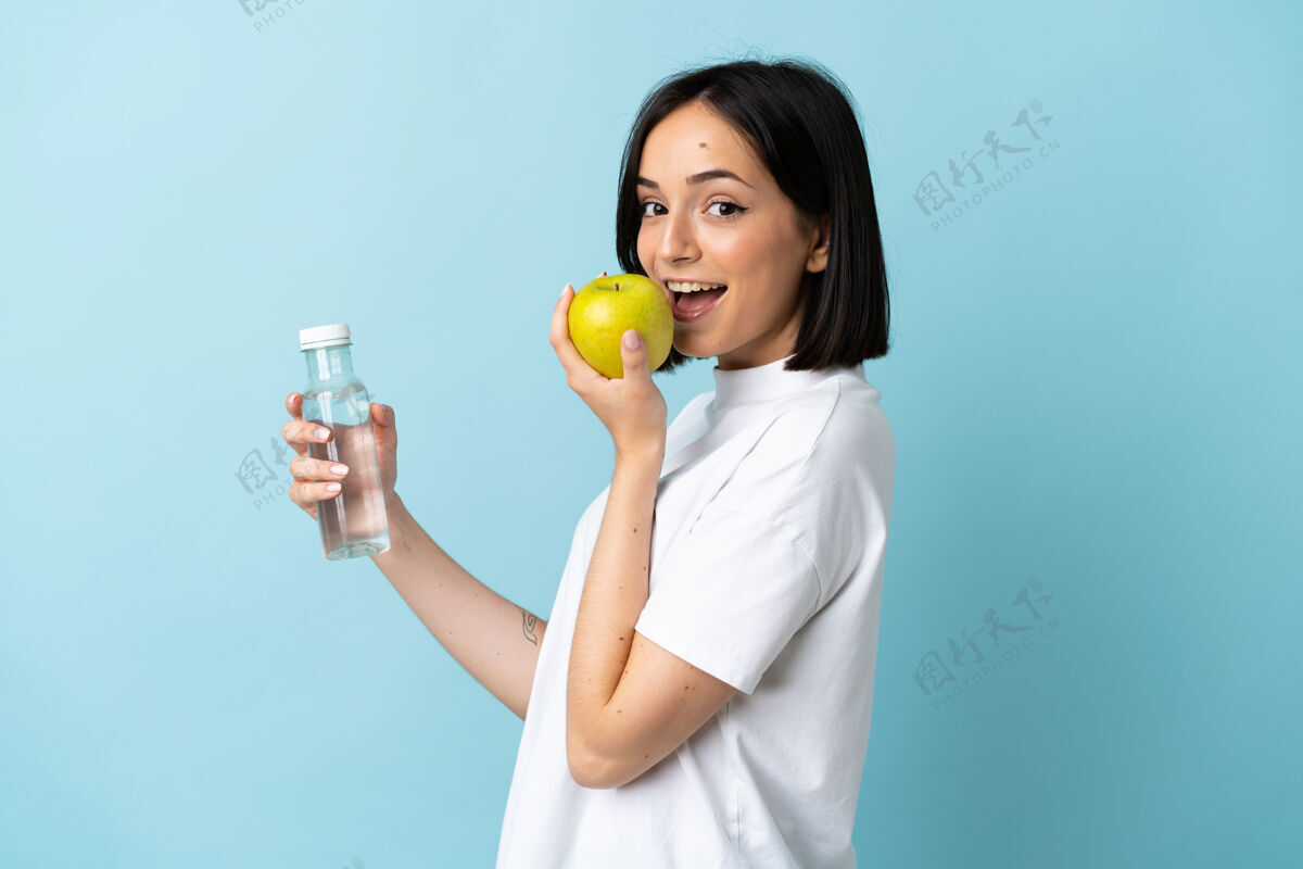 饮料一个年轻的白人妇女拿着一个苹果和一瓶水被隔离在蓝色的墙上新鲜矿物五颜六色
