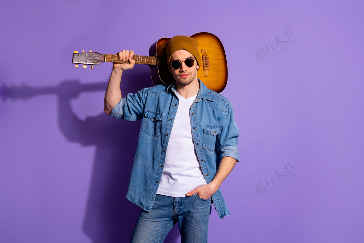 休闲照片中自信迷人的帅哥戴着棕色帽子眼镜 肩上扛着吉他 手放在口袋里 与紫色鲜艳的色彩背景隔离开来休息帽子吉他手