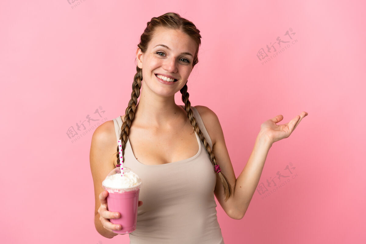 电话一位年轻的女士拿着草莓奶昔 孤立地站在粉红色的背景上 向旁边伸出双手 欢迎前来食物饮料杯子