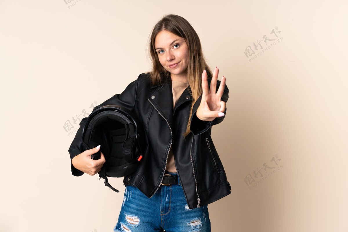 女性立陶宛年轻女子手持摩托车头盔 在米色背景下快乐地数着三个手指计数年轻女孩