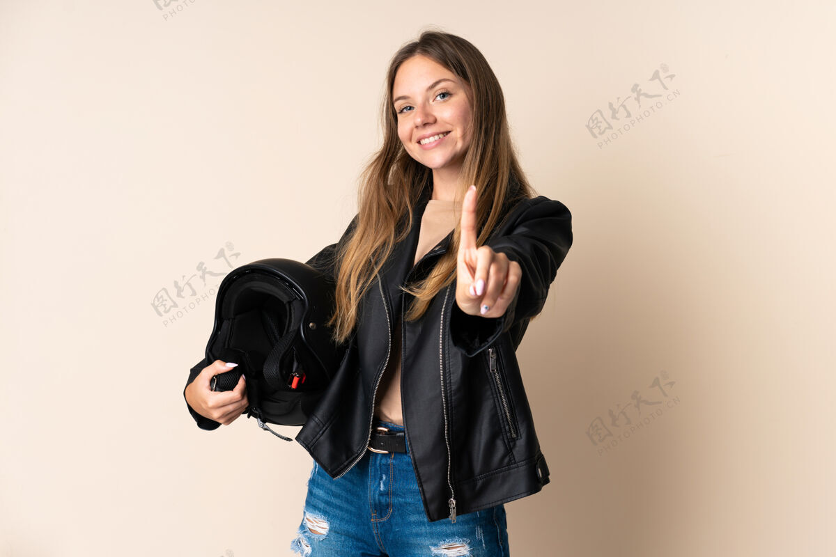 想法立陶宛年轻女子手持一个摩托车头盔 在米色背景下展示并举起手指皮革计数驾驶