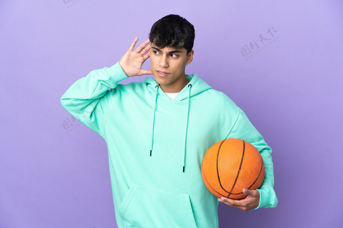 交流在孤立的紫色背景上打篮球的年轻人把手放在耳朵上听东西爱好听力减肥