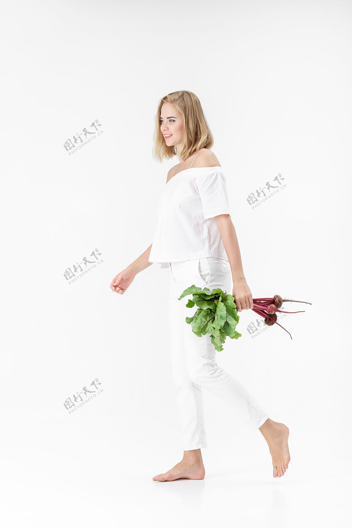 人一位穿着白色上衣的金发美女手持一根长着绿叶的甜菜根背景.健康还有维生素年轻背景夏天