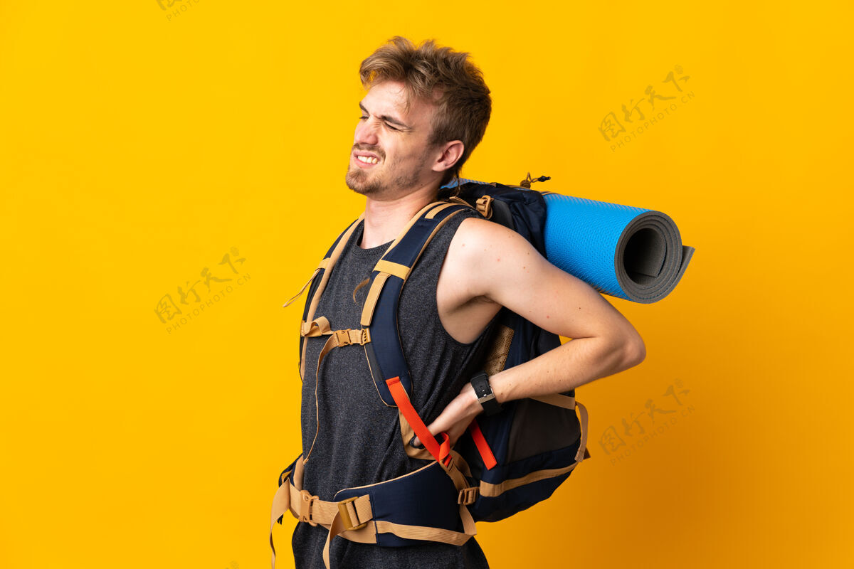 男性背着一个大背包的年轻登山者 被隔离在黄色的背景上 因为努力而背痛远足者旅游徒步旅行