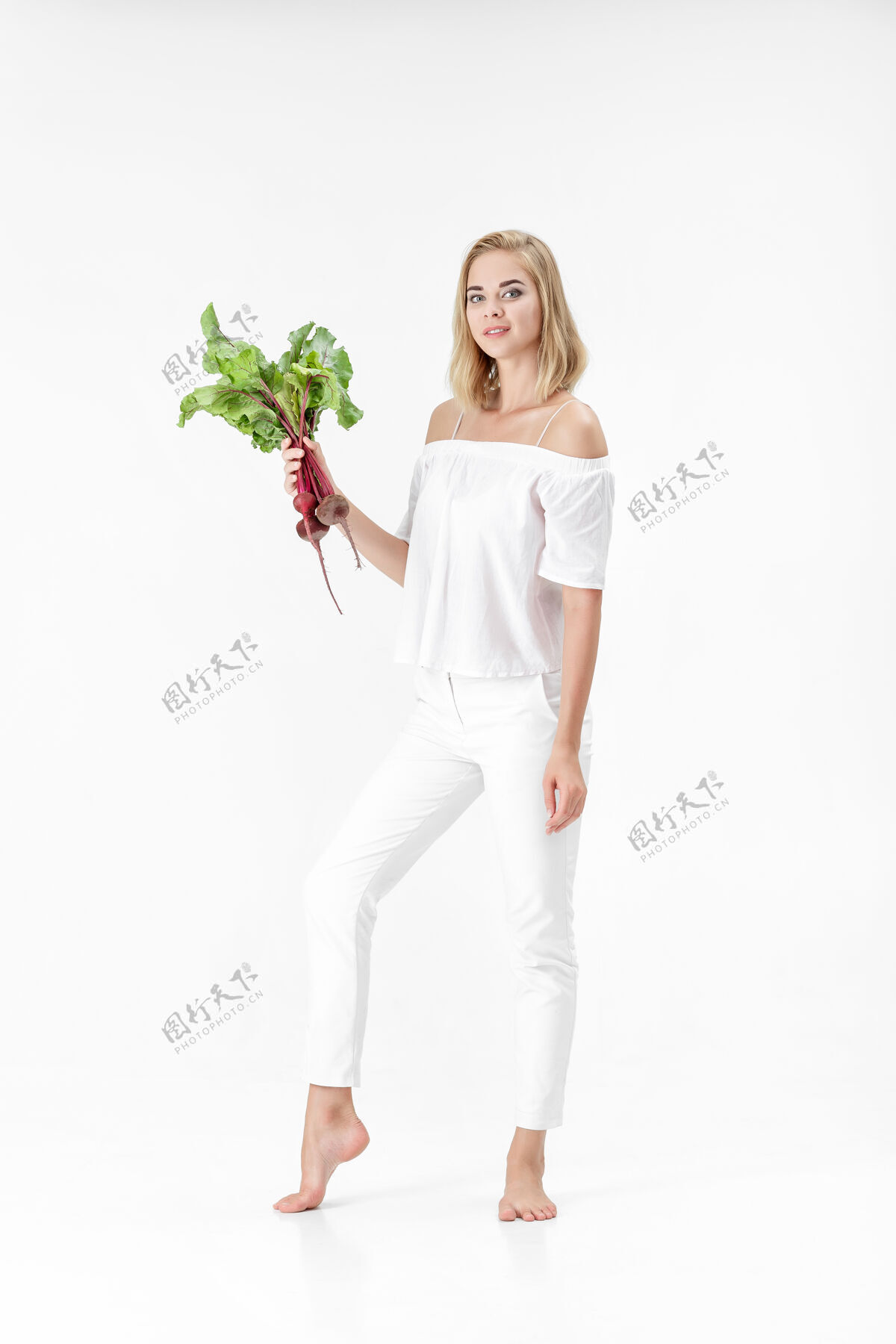 农场一位穿着白色上衣的金发美女手持一根长着绿叶的甜菜根背景.健康还有维生素快乐健康蔬菜
