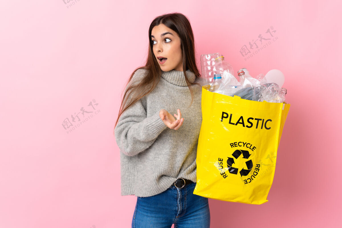环境年轻的白种女人拿着一个装满塑料瓶的袋子在粉红色的背景下 一边看着一边惊讶的表情责任年轻惊喜