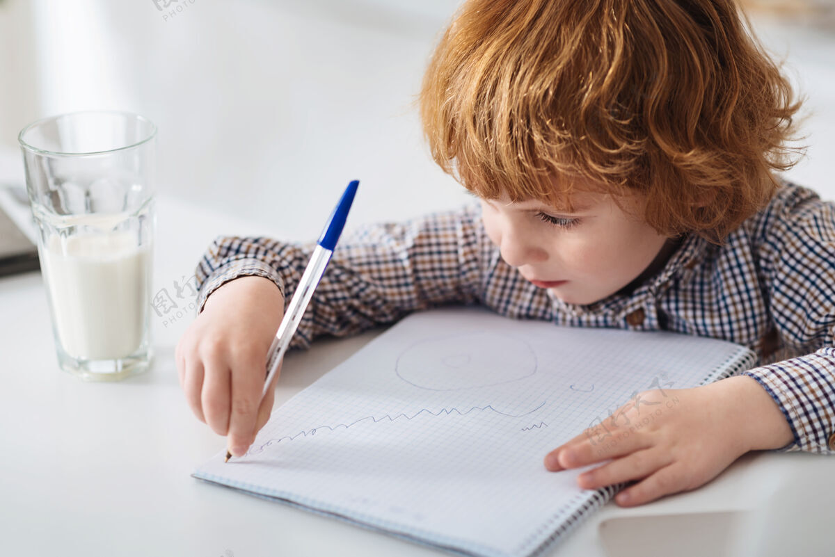 智力在一个杰作浓缩聪明可爱的孩子坐在白色的桌子旁 用钢笔在笔记本上画有趣的图画技术杯子早晨