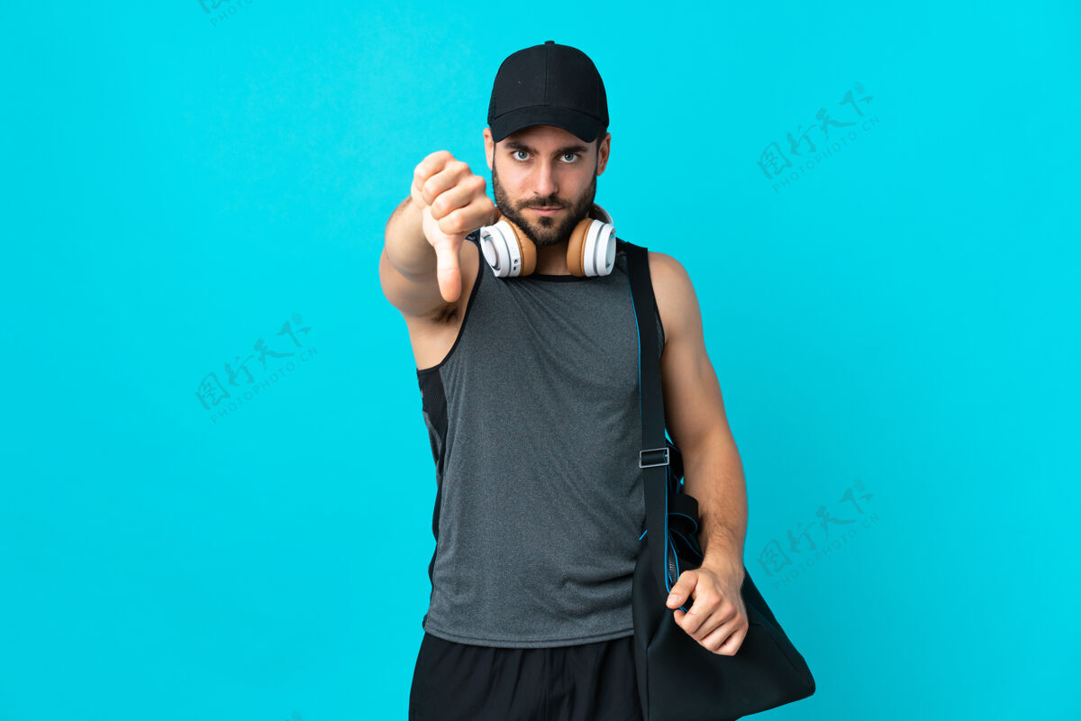 护理年轻的运动男子与运动包隔离在蓝色背景显示拇指向下与消极的表情健身房男性男人