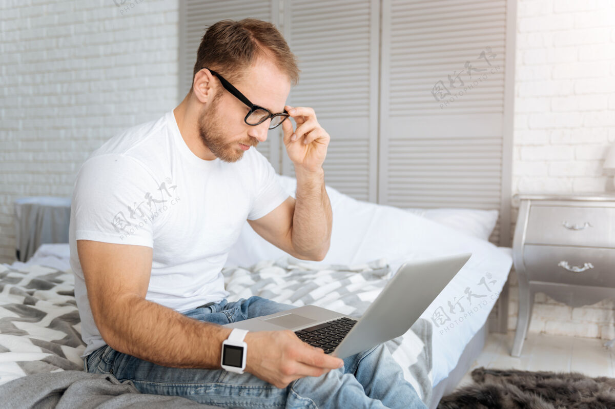 程序员时间到了工作快乐专注的大胡子男人坐在家里的床上 一边用笔记本电脑一边表达专注和工作科技当代电脑