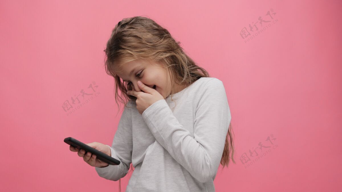 童年快乐可爱的小女孩手持智能手机 站在粉色背景上大笑的照片休闲兴趣活动