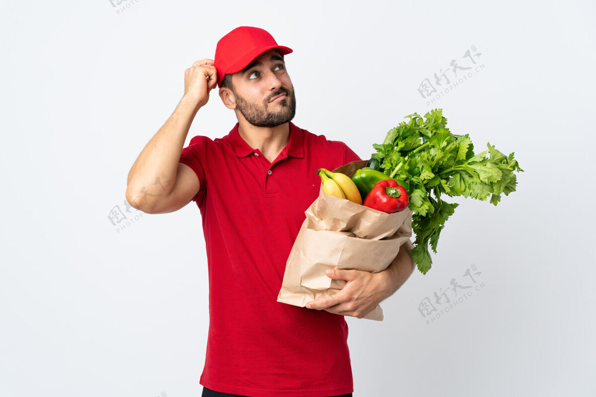 外卖一个留着胡子的年轻人拿着一袋蔬菜 被隔离在白色的背景上 脸上带着疑惑和迷惑的表情自然袋子成人