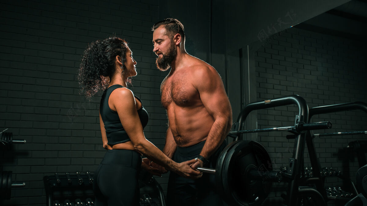 体重肌肉发达的赤膊男子和美丽的运动女子看着对方 同时举起沉重的杠铃在黑暗的健身房在一起健康关系训练