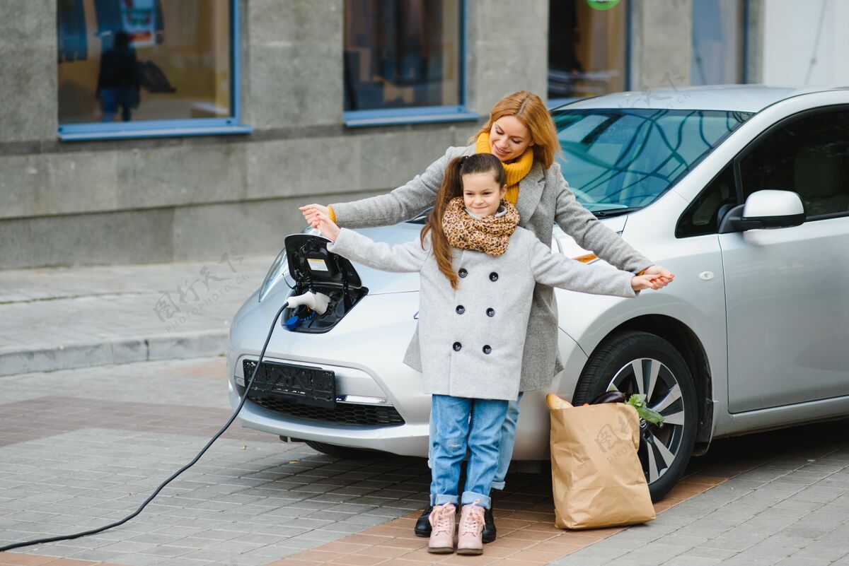 充电妈妈带着女儿在电动加油站给电动车充电 用手机说话电力选择休闲