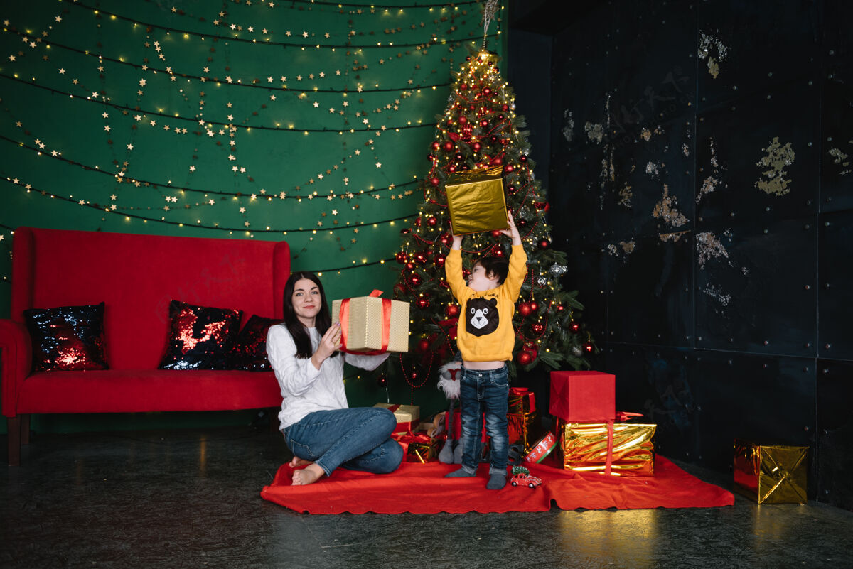 男孩年轻美丽的母亲带着可爱的儿子在圣诞树旁礼物快乐圣诞节童年父母圣诞树