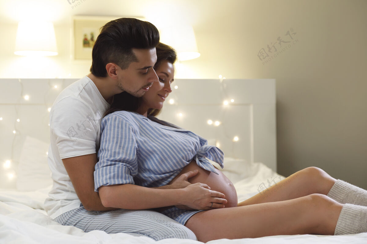 肚子一对年轻夫妇在等一场婚礼宝贝老公和他怀孕的妻子抱在一起坐在床上肥料放松睡觉时间