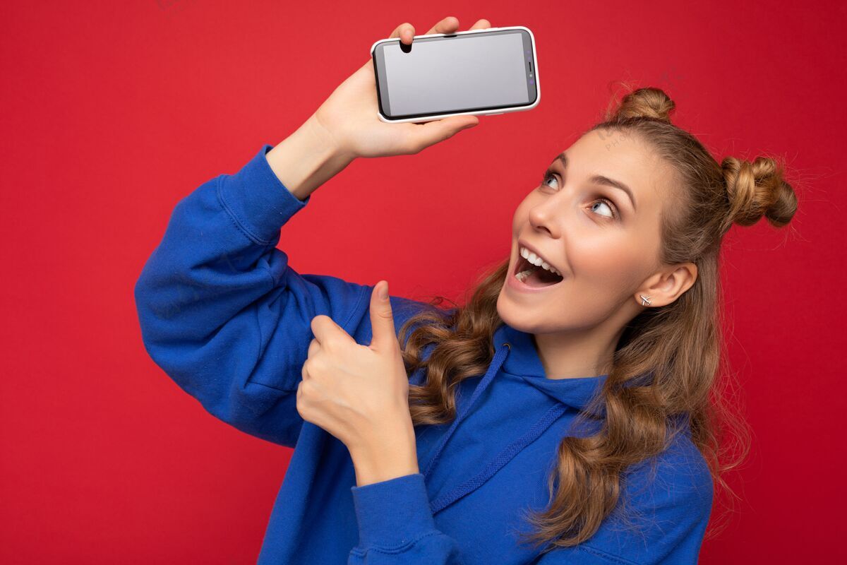 快乐迷人的震惊微笑的年轻金发女子穿着时尚的蓝色连帽衫隔离在红墙生动休闲手机